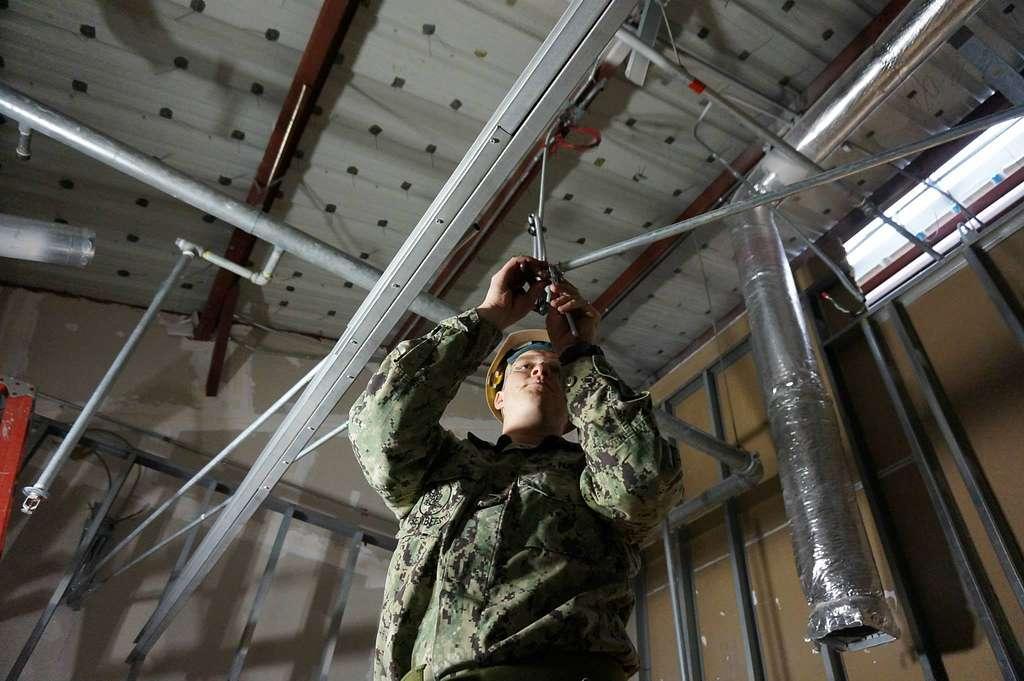 A U.S. Navy construction electrician fastening overhead metal conduit inside an unfinished building.